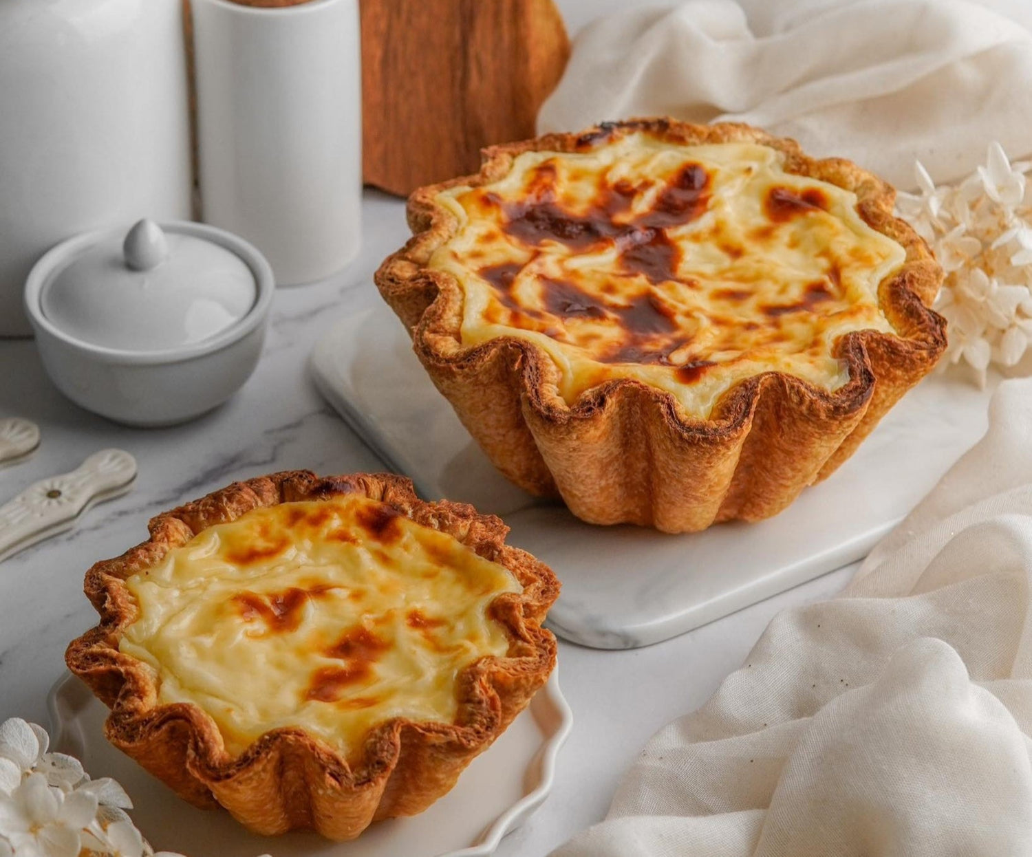 Two golden baked Buko Flan with caramelized tops in fluted pastry shells, served on white marble board with kitchen accessories.